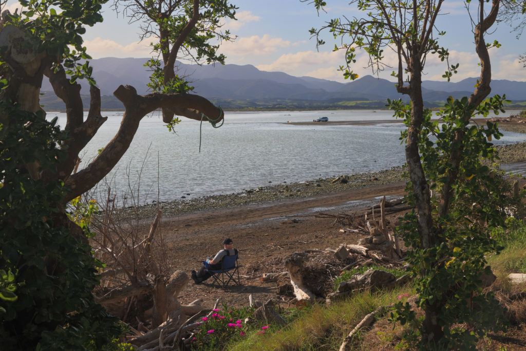 Image of Lake Ferry looking through the trees and a man sitting on the beach reclining on a char at dusk