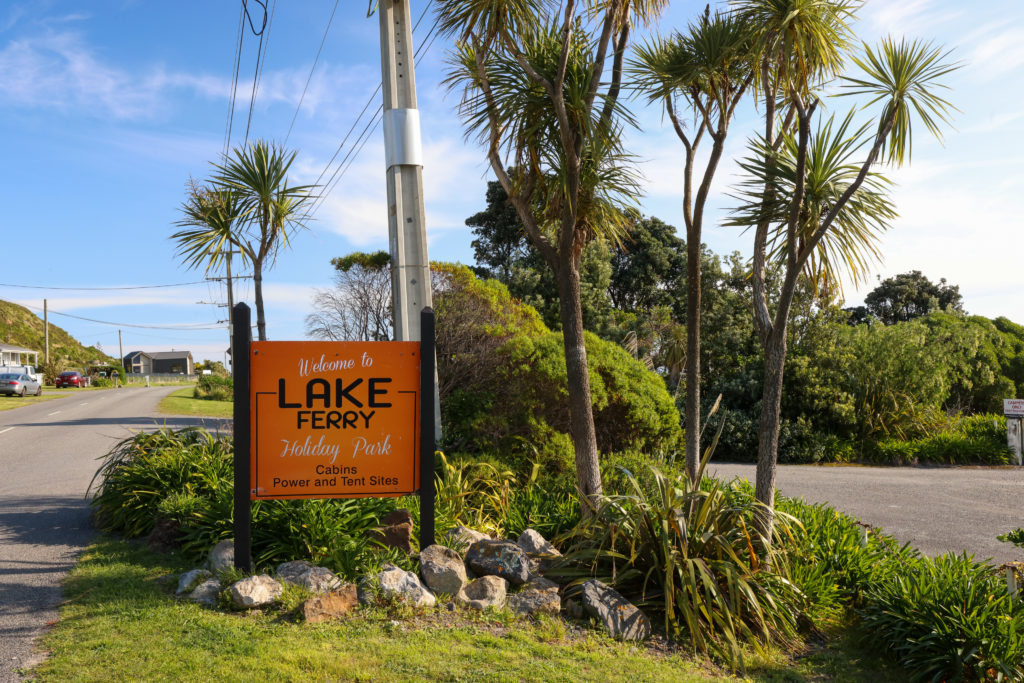 This image features an orange Welcome to Lake Ferry sign next to three cabbage trees